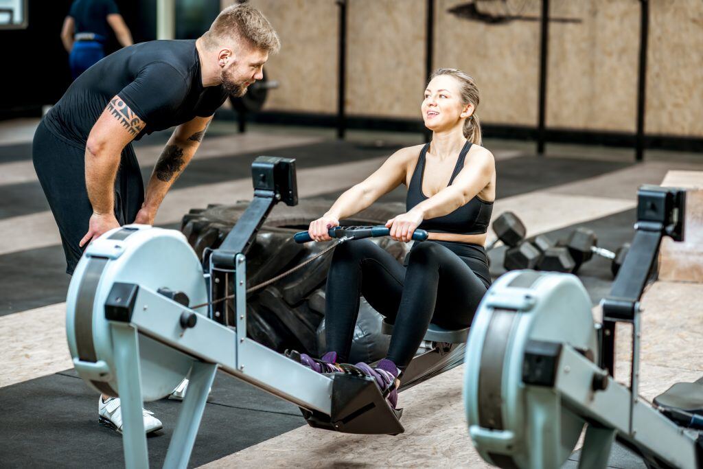 Attraktiver Trainer trainiert Frau am Rudergerät im Fitnessstudio - Wie auch beim Sport sollte auch beim Coaching für Führungskräfte und beim Business Coaching Der Coach nicht Wasser predigen und Wein trinken. Porsche wissen und praktische Erfahrung ist Essenziel.