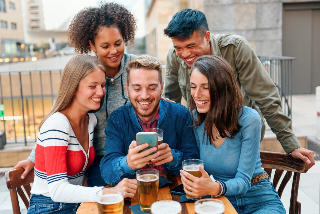 Eine Gruppe junger Menschen lacht und schaut auf ein Handy, Recruiting über Social Media ist längst zur Pflicht geworden