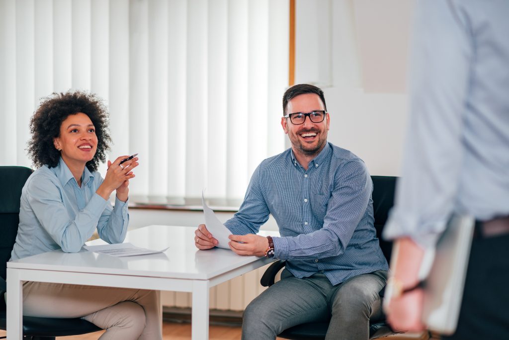 2 Mitarbeiter lachen im Pausenraum: Wenn Sie auf der Suche nach Recruiting Ideen sind, stellen Sie unbedingt Mitarbeiter in den Mittelpunkt ihrer HR Kampagne.