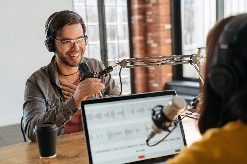  Zwei Podcaster mit Kopfhörern lachen, reden und nehmen einen Live-Podcast auf. - B2B Podcast Marketing Ist eine lukrative Methode, um Content zu generieren Und die Markenbekanntheit zu stärken